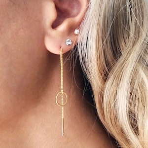 May include: A gold chain earring with a small circle charm hanging from it. The earring is worn in the lower lobe of the ear.