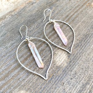 Puede incluir: Un par de pendientes de aro de plata con un colgante de cristal rosa que cuelga de cada aro. Los aros tienen forma de hoja y los cristales son iridiscentes.