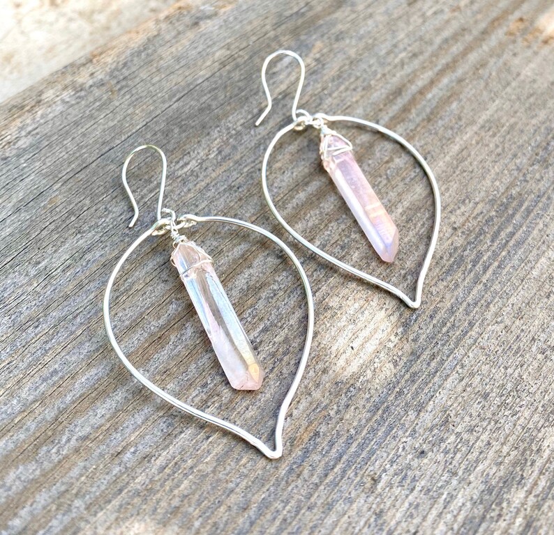 Puede incluir: Un par de pendientes de aro de plata con un colgante de cristal rosa que cuelga de cada aro. Los aros tienen forma de hoja y los cristales son iridiscentes.
