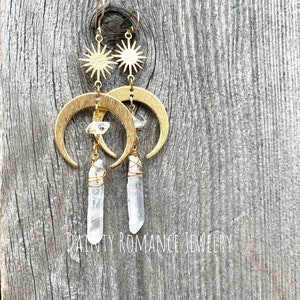 Pu&ograve; includere: Orecchini a forma di mezzaluna dorati con cristalli di quarzo trasparenti e ciondoli a forma di stella. Gli orecchini sono appesi a una superficie di legno.