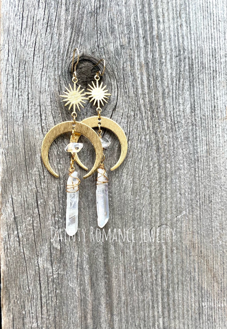 Pu&ograve; includere: Orecchini a forma di mezzaluna dorati con cristalli di quarzo trasparenti e ciondoli a forma di stella. Gli orecchini sono appesi a una superficie di legno.