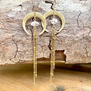 May include: Gold crescent moon earrings with a clear crystal charm and multiple gold chains dangling from the bottom.