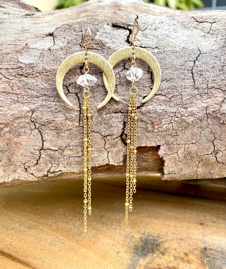 May include: Gold crescent moon earrings with a clear crystal charm and multiple gold chains dangling from the bottom.