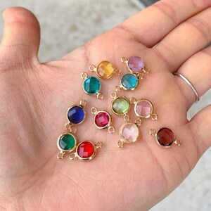 May include: Assortment of 12 round glass connector charms with gold-toned metal accents. The charms are in various colors, including red, blue, green, pink, and clear.