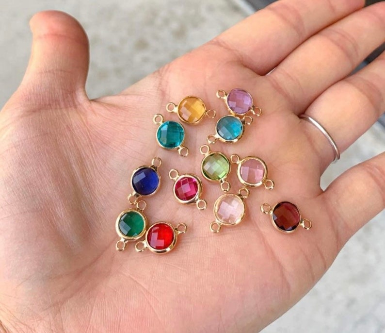 May include: Assortment of 12 round glass connector charms with gold-toned metal accents. The charms are in various colors, including red, blue, green, pink, and clear.