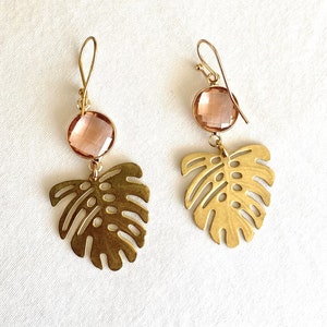 May include: A pair of gold earrings with a pink crystal and a cutout monstera leaf design.