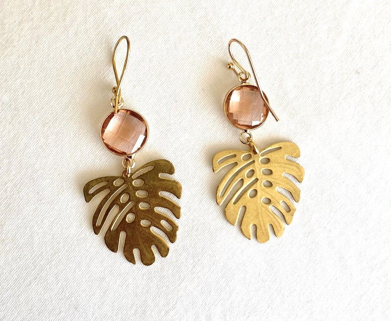 May include: A pair of gold earrings with a pink crystal and a cutout monstera leaf design.