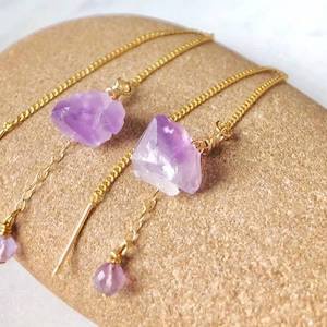 Puede incluir: Tres pares de pendientes de cristal de amatista con cadenas doradas y pequeñas gotas de amatista. Los pendientes se muestran sobre una piedra marrón claro.