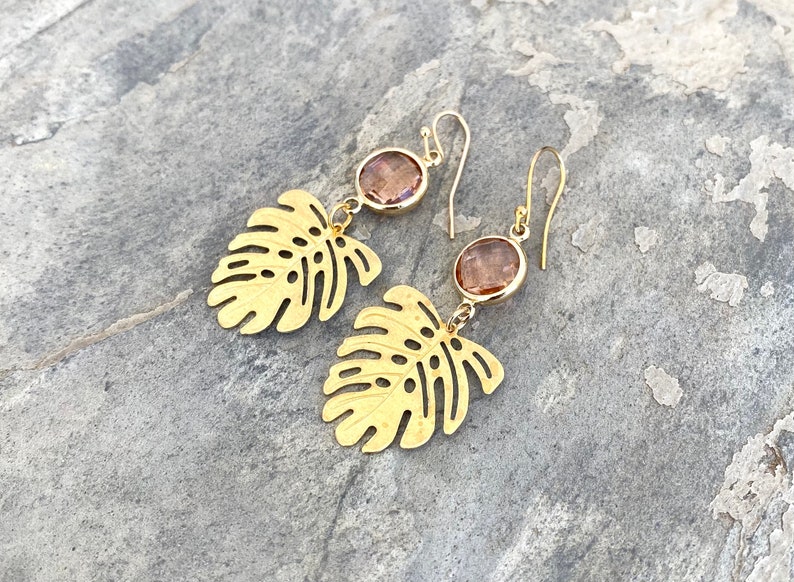 May include: Gold earrings with a leaf design and a pink gemstone dangle. The earrings are made of metal and have a delicate, feminine look.