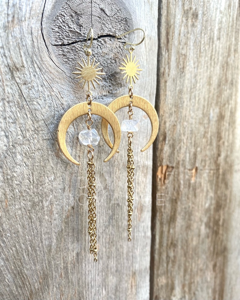 May include: Gold crescent moon earrings with a small clear crystal and a gold sun charm dangling from each earring. The earrings have a delicate gold chain that hangs below the crystal.