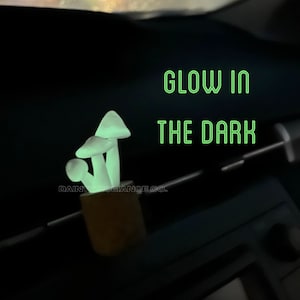 Puede incluir: Un conjunto de tres ambientadores de coche en forma de hongo blanco que brillan en la oscuridad. Los hongos están en una pequeña maceta marrón. El texto "Glow in the Dark" está escrito en letras verdes sobre un fondo negro.