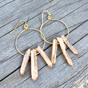 May include: Gold hoop earrings with a cluster of light brown crystal points dangling from each hoop.