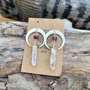 Pu&ograve; includere: Orecchini a forma di mezzaluna d'argento con perline di ametista e cristalli di quarzo trasparenti che pendono da essi. Gli orecchini sono esposti su una carta marrone con il testo "Dainty Roman Jewelry".