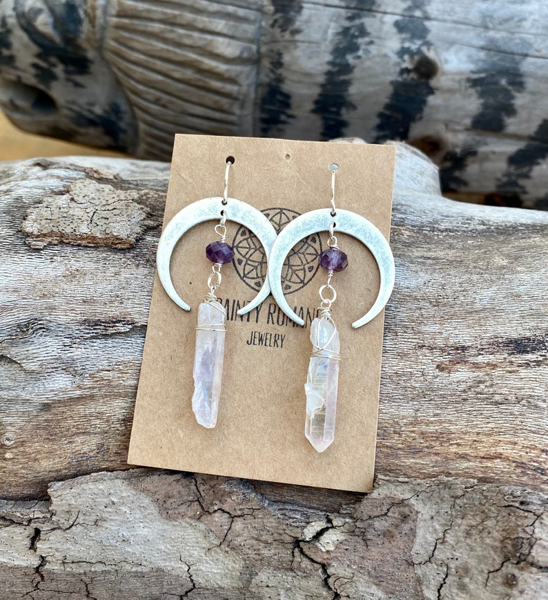 Pu&ograve; includere: Orecchini a forma di mezzaluna d'argento con perline di ametista e cristalli di quarzo trasparenti che pendono da essi. Gli orecchini sono esposti su una carta marrone con il testo "Dainty Roman Jewelry".