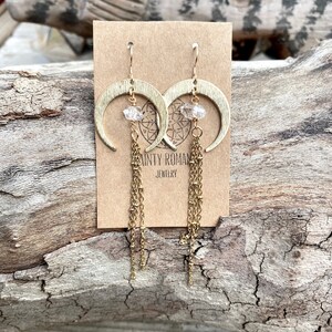 May include: Gold crescent moon earrings with clear crystal accents and dangling gold chains. The earrings are displayed on a brown card with the text "Sainty Roman Jewelry".