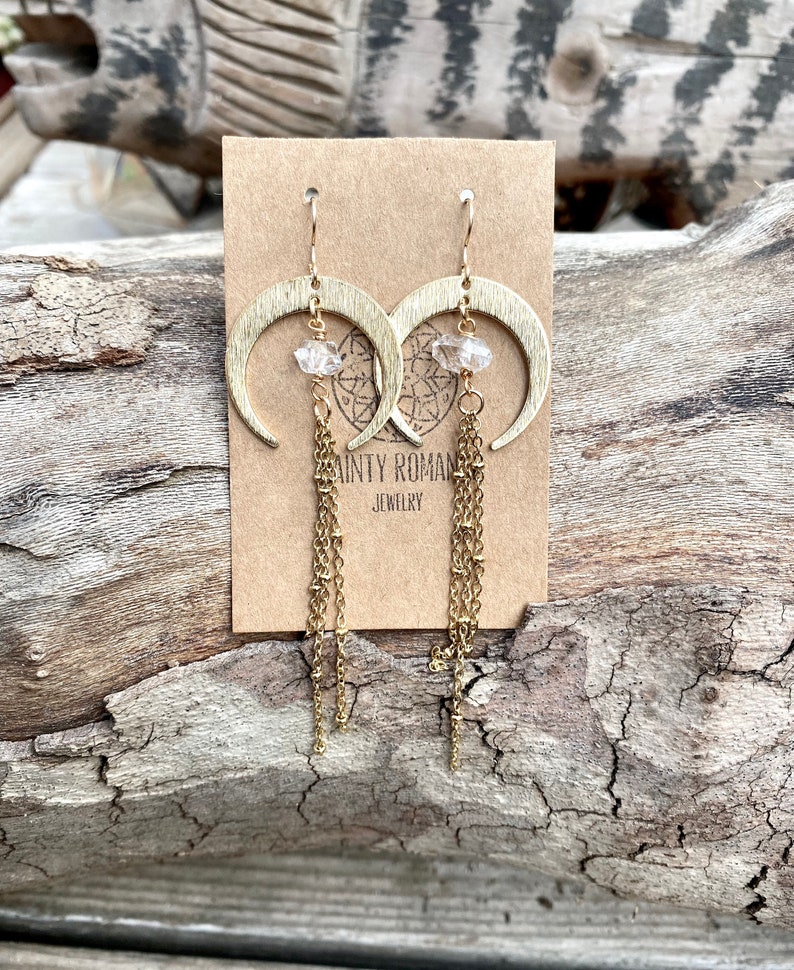 May include: Gold crescent moon earrings with clear crystal accents and dangling gold chains. The earrings are displayed on a brown card with the text "Sainty Roman Jewelry".