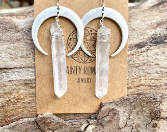 May include: Silver-tone crescent moon earrings with clear quartz crystal pendants. The crystals are wrapped in silver wire and hang from the moon-shaped hoops. The earrings are displayed on a brown card with the text "DAINTY ROMANCE JEWELRY".