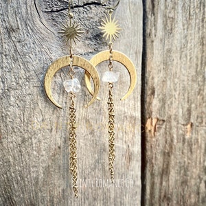 May include: Gold crescent moon earrings with a clear crystal charm and a star charm dangling from the top. The earrings have a delicate chain that hangs below the crystal charm. The earrings are photographed against a wooden background.