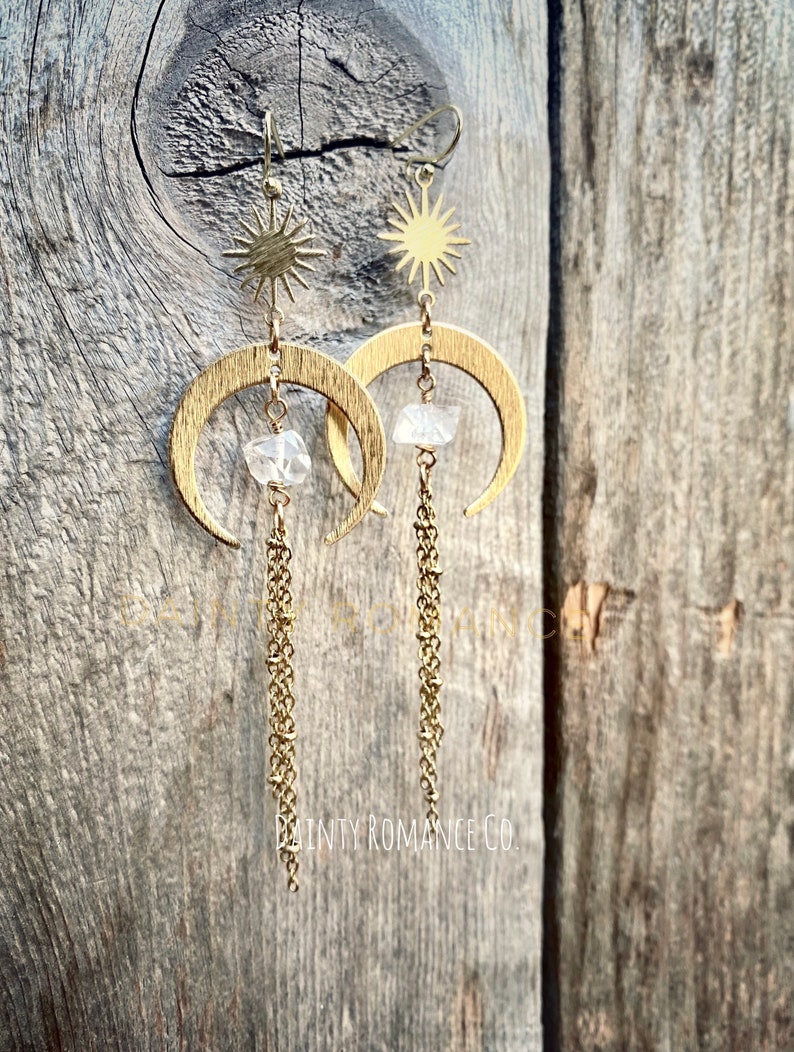 May include: Gold crescent moon earrings with a clear crystal charm and a star charm dangling from the top. The earrings have a delicate chain that hangs below the crystal charm. The earrings are photographed against a wooden background.