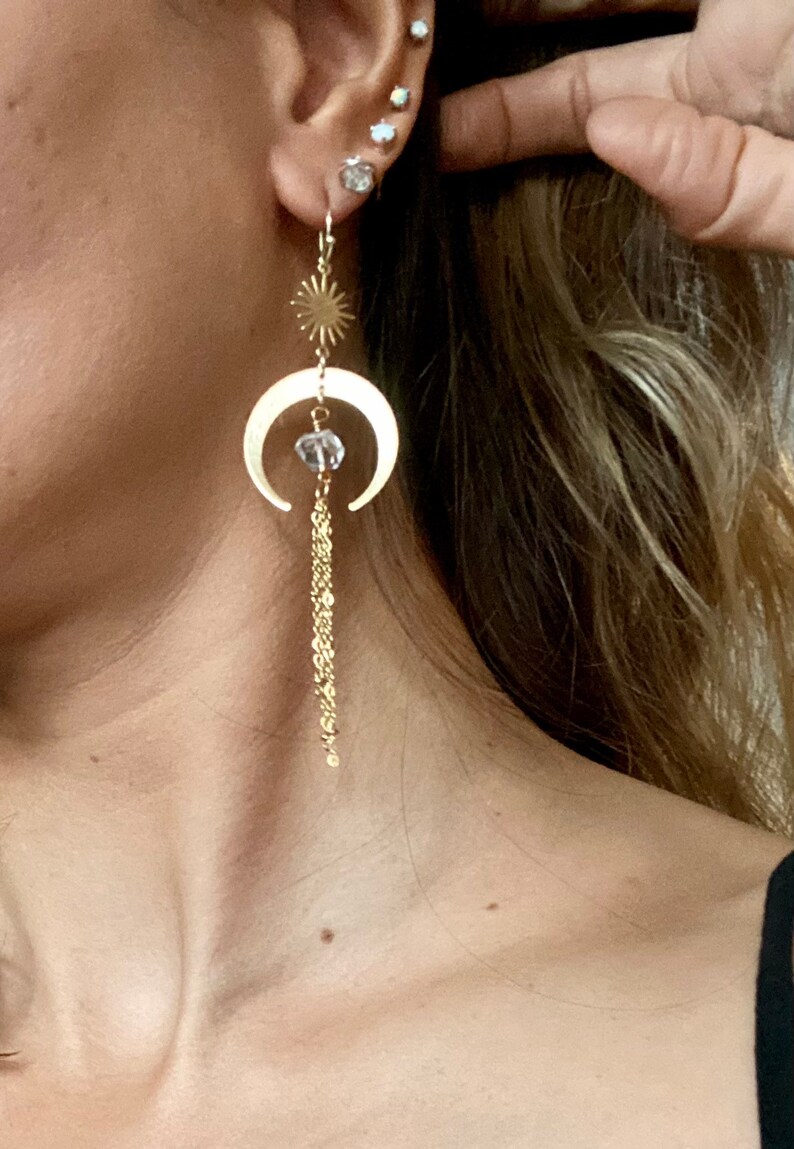 May include: Gold crescent moon earrings with a small crystal bead and a dangling chain. The earrings feature a sun charm at the top.