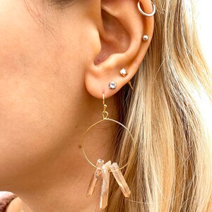 May include: Gold hoop earrings with a cluster of light brown crystal points dangling from the bottom.