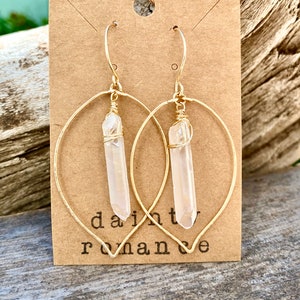 Puede incluir: Aretes de aro dorados con un colgante de cristal de cuarzo transparente. Los aretes est&aacute;n en una tarjeta marr&oacute;n con el texto "dainty romance".