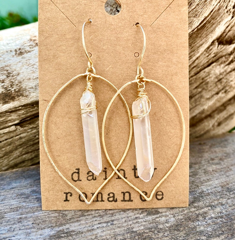 Puede incluir: Aretes de aro dorados con un colgante de cristal de cuarzo transparente. Los aretes est&aacute;n en una tarjeta marr&oacute;n con el texto "dainty romance".