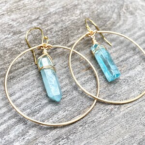 Puede incluir: Aretes de aro de oro con un colgante de cristal azul claro que cuelga de cada aro.