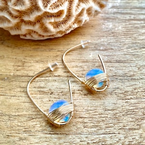 May include: Gold hoop earrings with a light blue opal gemstone wrapped in gold wire.
