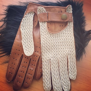 Op de afbeelding: Een paar bruine leren rijhandschoenen met een beige gebreide rug. De handschoenen hebben een band met een knoopsluiting aan de achterkant van de hand.