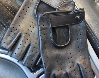 Puede incluir: Un par de guantes de conducir de cuero negro con diseño sin dedos y detalles perforados. Los guantes tienen una correa de muñeca con cierre de presión.