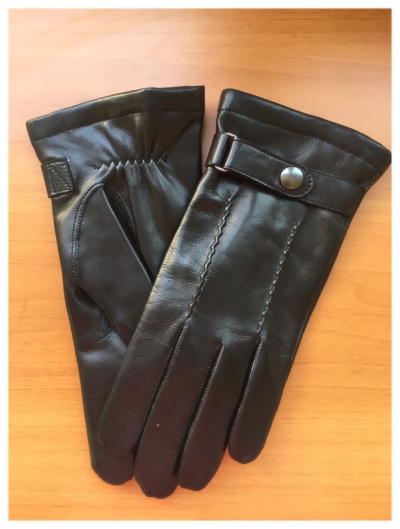 Op de afbeelding: Een paar zwarte leren handschoenen. De handschoenen hebben een gestikt ontwerp en een bandje met een zilveren knoopsluiting. De handschoenen zijn gemaakt van glad, glanzend leer.