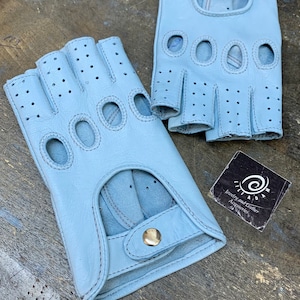 Può includere: Un paio di guanti da guida in pelle blu chiaro con fori per le dita e chiusura a scatto. I guanti sono in pelle traforata e hanno un colore blu chiaro.