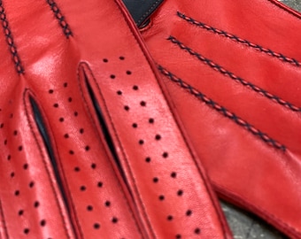 Puede incluir: Guantes de conducción de cuero rojo con detalles negros y perforaciones. Los guantes tienen costuras negras y un cierre a presión. La imagen también muestra dos cierres a presión a juego y una tarjeta de visita con un logotipo.