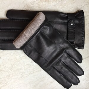 Op de afbeelding: Een paar zwarte leren handschoenen met een lichtbruine gebreide manchet. De handschoenen hebben een gestikt ontwerp en een bandje met een zilveren knoopsluiting. De handschoenen liggen op een lichtgekleurd oppervlak.