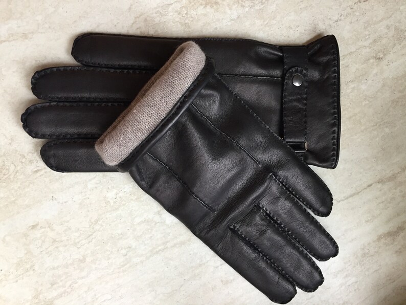 Op de afbeelding: Een paar zwarte leren handschoenen met een lichtbruine gebreide manchet. De handschoenen hebben een gestikt ontwerp en een bandje met een zilveren knoopsluiting. De handschoenen liggen op een lichtgekleurd oppervlak.