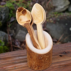 Puede incluir: Dos cucharas de madera descansan en un pequeño y rústico soporte de madera. Una cuchara tiene un color de madera claro, mientras que la otra tiene una veta de madera más oscura y estampada. El soporte es cilíndrico con un exterior de corteza natural.