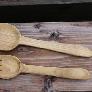 May include: Two wooden salad utensils: a large spoon and a fork with three tines. The light-colored wood grain is visible on both pieces. The utensils are displayed on a wooden surface.