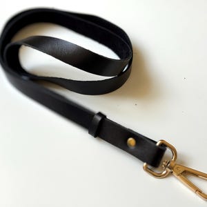 Puede incluir: Un cordón de cuero negro con un acabado mate liso y un mosquetón de metal dorado. La correa está asegurada con una trabilla de cuero negro a juego y un pequeño remache dorado, creando un accesorio funcional para sujetar objetos.