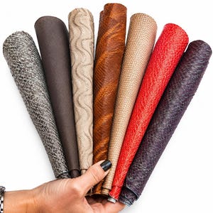 Op de afbeelding: Een waaier van zeven opgerolde stukken leer in verschillende kleuren en texturen, waaronder grijs slangenleer, donkerbruin, beige met een golvend patroon, oker, lichtbeige slangenleer, felrood en donkerpaars met een slangenleerstructuur.