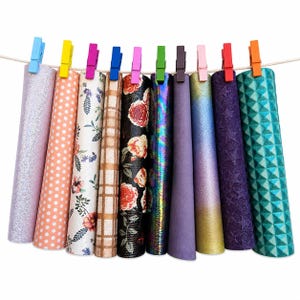 Puede incluir: Diez rollos verticales de material para manualidades con diversos patrones y texturas, incluyendo purpurina, lunares, flores, cuadros, rosas, iridiscente, morado liso, degradado, estrellas en relieve y geométrico, sujetos por pinzas.