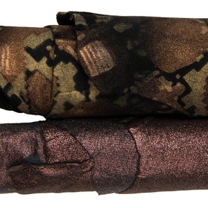 Op de afbeelding: Twee rollen stof liggen horizontaal op elkaar gestapeld. De bovenste rol heeft een donkerbruin en olijfgroen camouflagepatroon met een metallic glans. De onderste rol is effen, diep metallic brons met een getextureerd, licht gekreukt oppervlak.