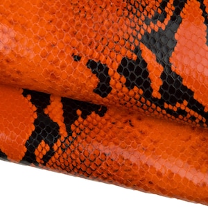 May include: Close-up of orange and black snakeskin leather. The leather has a textured, scaly pattern with a vibrant orange base and black markings. The material is folded, revealing the underside and adding depth.