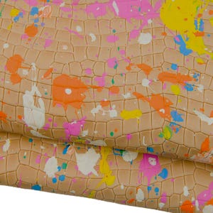May include: Close-up of a textured, tan surface with a pattern of irregular shapes, resembling reptile skin. The surface is splattered with colourful paint in shades of pink, orange, yellow, blue, and white, creating a vibrant, artistic design.