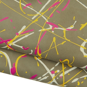 May include: Close-up of a fabric with a taupe background, overlaid with abstract paint splatters in white, yellow, and hot pink. The fabric is folded, revealing its texture and the layered design.