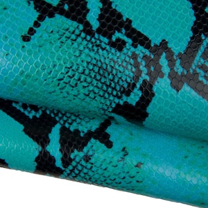 Puede incluir: Primer plano de un patrón de piel de serpiente turquesa y negro. El patrón presenta un diseño texturizado, similar a escamas, con un patrón repetitivo de formas negras sobre un fondo turquesa brillante. La imagen muestra el material doblado.