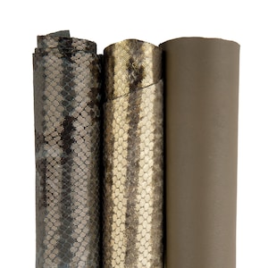 Może przedstawiać: Trzy rolki tkaniny, jedna z wzorem skóry węża w odcieniach szarości i złota, druga z wzorem skóry węża w odcieniach złota i bieli oraz jedna w jednolitym kolorze szarym.