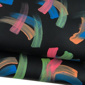 Op de afbeelding: Close-up van zwart materiaal met kleurrijke penseelstreken. De streken zijn in roze, oranje, groen en blauw, waardoor een levendig abstract patroon ontstaat. Het materiaal lijkt gevouwen, waardoor de textuur en diepte van het ontwerp zichtbaar worden.