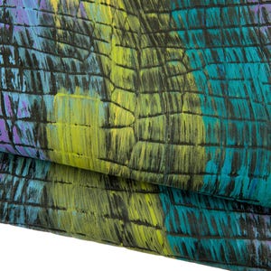 Op de afbeelding: Close-up van een textiel met een patroon dat op reptielenhuid lijkt. Het ontwerp bevat een mix van kleuren, waaronder teal, geel en paars, op een zwarte achtergrond. De stof is gevouwen, waardoor de gelaagde textuur zichtbaar wordt.