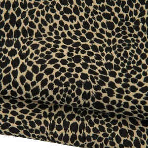 Puede incluir: Una pieza de tela doblada con un patrón repetitivo de manchas de leopardo negras sobre un fondo de color tostado claro. El material muestra pliegues y dobleces suaves, resaltando la textura y las manchas densas e irregulares.